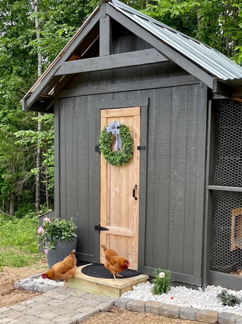 Puede incluir: Un gallinero de madera gris con una puerta de madera y una corona verde. Dos gallinas marrones est&aacute;n frente al gallinero.