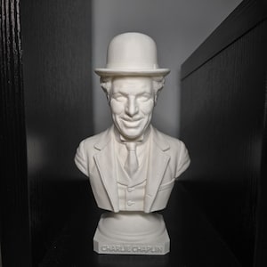 Pode incluir: Um busto branco de Charlie Chaplin, com seu chapéu coco e terno. O busto está montado em um pedestal com a inscrição "CHARLIE CHAPLIN". A escultura é exibida em uma prateleira preta.