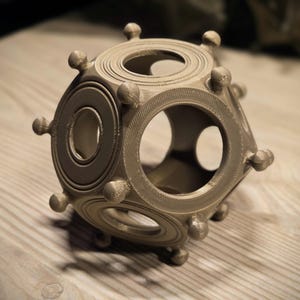 Può includere: Un oggetto geometrico color beige, stampato in 3D, con aperture circolari e ovali. L'oggetto presenta un design complesso con più anelli circolari e piccole sporgenze arrotondate. È appoggiato su una superficie di legno chiaro.