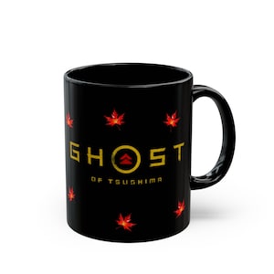 Puede incluir: Taza de cerámica negra con el texto dorado "GHOST OF TSUSHIMA", un emblema circular y diseños de hojas de arce rojas. La taza tiene un asa negra y un acabado brillante.