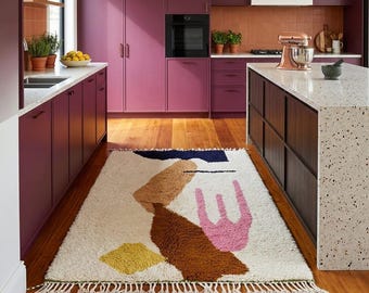 May include: A cream-colored, rectangular rug with an abstract design in shades of brown, pink, yellow, and navy blue. The rug has a fringed edge and is placed on a wooden floor in a kitchen setting. The kitchen features magenta cabinets and a white countertop.