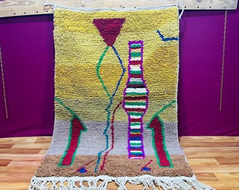 May include: A colorful, abstract rug with a yellow, brown, and gray background. The rug features geometric shapes in red, green, and blue, including arrows and a bottle-like design. The rug is approximately 48 inches wide and 72 inches long.