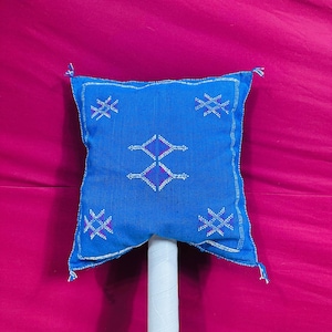Op de afbeelding: Een vierkant, blauw decoratief kussen met een geometrisch ontwerp. Het kussen heeft een centraal ruitpatroon en stervormige vormen, allemaal in wit en paars. Het kussen heeft kwastjes op elke hoek.