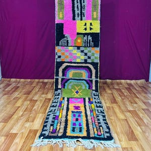 May include: A vibrant, rectangular rug with a geometric pattern. The rug incorporates pink, black, green, yellow, and blue hues. It has a fringe along the bottom edge and is shown on a wooden floor. The rug's design is intricate.