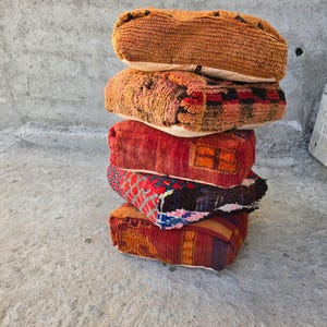 May include: A vertical stack of five rectangular floor cushions featuring varied woven textures and patterns. The cushions display a palette of orange, red, black, and blue tones with intricate geometric designs and frayed edges on several pieces.