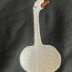 May include: A black garment featuring an embroidered goose design. The goose has a white body, a long neck, and an orange beak and feet. The embroidery is detailed and the goose is centered on the garment.
