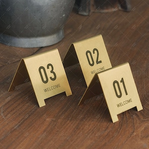 May include: Three gold-coloured metal table numbers, numbered 01, 02, and 03, with the word "WELCOME" printed below each number. The table numbers are angled and stand on a wooden surface.