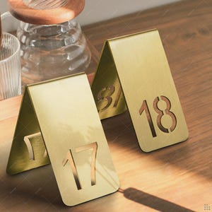 May include: Three gold-colored table number holders with cut-out numbers 17, 18, and 31. The holders are angled, creating a triangular shape. The numbers are in a sans-serif font. The background is a wooden surface.