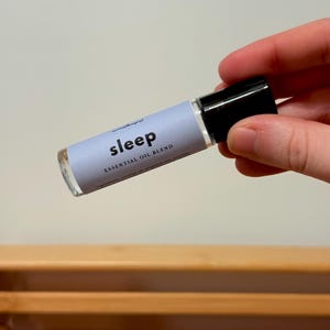 May include: A clear glass essential oil roller bottle with a black cap. The label reads "sleep" in a modern font, with "essential oil blend" below. The bottle is held against a neutral background.