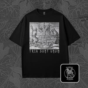 May include: Black t-shirt featuring a detailed black and white graphic with the text "TRIA SUNT UNUM." The graphic depicts three figures and a serpent. A small square logo with a winged lion and the words "Nocturnal Academy" is in the lower right corner.