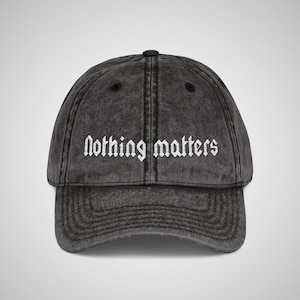 May include: A charcoal grey acid-wash baseball cap featuring the text Nothing matters embroidered in white gothic-style lettering across the front panels. The cap has a curved brim, a button on top, and visible stitching along the seams.