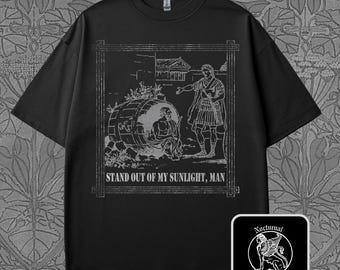 May include: Black t-shirt with a gray graphic depicting a classical scene with a man in a barrel and another gesturing. The text "STAND OUT OF MY SUNLIGHT, MAN" is below the image. A small square logo with a winged lion is in the lower right.