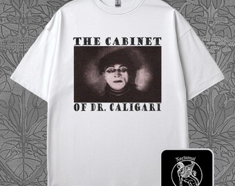 May include: White t-shirt featuring a black and white image from the film "The Cabinet of Dr. Caligari." The text "THE CABINET OF DR. CALIGARI" is printed above and below the image. A small, square logo with a winged creature is in the lower right corner.