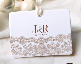 Peut inclure: Un ornement rectangulaire en céramique blanche avec un motif de dentelle crème sur le bord inférieur. Le centre affiche le texte marron J&R au-dessus de la date 10.10.2026. Un ruban crème est passé dans un trou en haut.