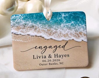 Peut inclure: Un ornement rectangulaire représentant une plage avec des vagues turquoise s'écrasant sur le sable beige. Le texte indique engaged, Livia & Hayes, 20.06.2026, et Outer Banks, NC. Un ruban blanc est fixé en haut pour suspendre.