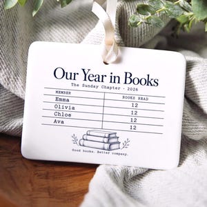 Peut inclure: Un ornement rectangulaire blanc en c&eacute;ramique avec un ruban cr&egrave;me, portant le texte noir Our Year in Books, The Sunday Chapter 2026, une liste de noms avec des nombres de livres, une illustration de trois livres et Good books. Better company.