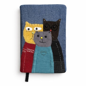 May include: A denim notebook with an appliqué design of three cartoon cats. The cats are yellow, grey, and black, each with unique features and clothing. The notebook has a black ribbon bookmark.