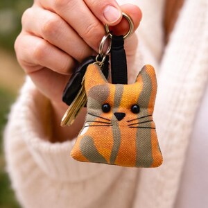 May include: A cat-shaped keychain with a black strap and gold ring. The cat is orange and green with black eyes and whiskers. The keychain is attached to keys.