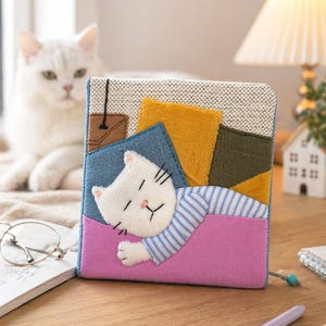 Puede incluir: Un artículo de tela cuadrado hecho a mano con un gato blanco durmiendo con pijama a rayas azules y una manta rosa. El fondo tiene parches de mezclilla, amarillo mostaza y verde oliva. Se ve un gato blanco.