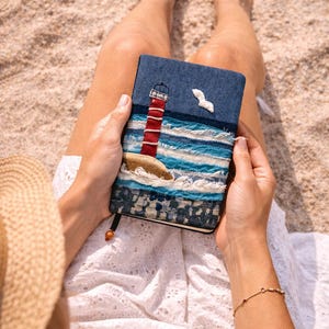 Puede incluir: Un diario de mezclilla con un diseño de faro y gaviota. La portada presenta un faro a rayas rojas y blancas, olas azules y una gaviota blanca. El diario se sostiene en una playa de arena.
