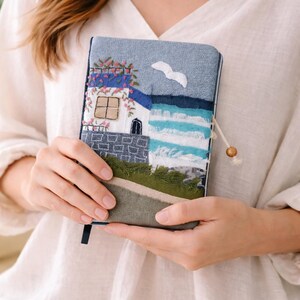 May include: A handmade journal with a coastal scene embroidered on the cover. The design features a white house with a blue roof, a seagull, and ocean waves. The journal has a blue ribbon bookmark and a wooden bead closure.