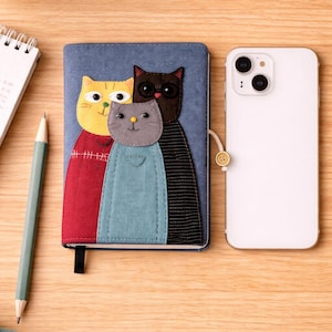 以下が含まれることがあります： 木製の表面に、猫のデザインの青いノート、白いスマートフォン、鉛筆、小さなメモ帳があります。ノートには、黄色、黒、グレーの3匹の猫の顔と、赤と青の衣装が描かれています。黒いブックマークが付いています。