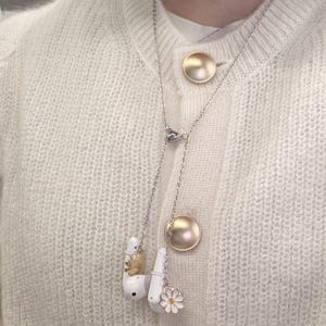 Peut inclure: Un collier argenté avec des écouteurs blancs, un pendentif doré et un charm en forme de marguerite. Le collier est superposé, avec un bouton doré sur le cardigan. Les écouteurs ont des détails dorés et blancs.