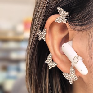 Puede incluir: Cierres de oreja de mariposa plateados con cristales transparentes brillantes. Los cierres están diseñados para colocarse en la oreja, creando la ilusión de mariposas. También se ve un auricular blanco.