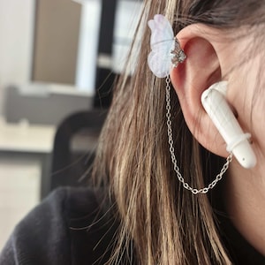May include: A delicate ear cuff featuring a butterfly design in shades of white and lavender, connected by a silver chain to a white earbud. The butterfly cuff is attached to the ear, adding a touch of whimsy to the earbud.