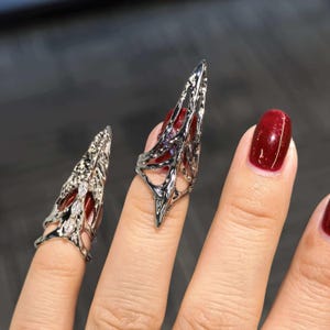 以下が含まれることがあります： 複雑な透かし彫りのデザインが施された、シルバーカラーのネイルリング。指に装着し、ドラマチックでゴシックな美しさを演出します。爪は深紅に塗られており、シルバーのリングを引き立てています。