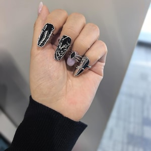 May include: A hand displaying several silver-toned rings with black enamel accents. The rings feature elongated, nail-shaped designs with intricate patterns and a pointed claw-like ring. The rings are a unique and edgy fashion accessory.