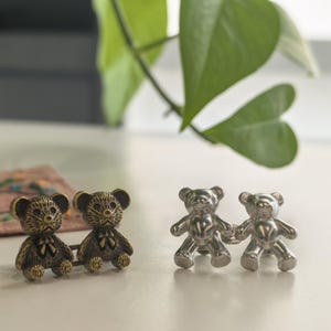 May include: Two pairs of bear-shaped cufflinks. One pair is bronze-colored, and the other is silver. The bear cufflinks are standing on a white surface. Green leaves are in the background.