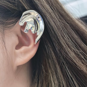 May include: A polished silver-tone ear cuff with a smooth, curved, and abstract organic shape that wraps around the upper cartilage of the ear. The metallic surface reflects light, highlighting the fluid, molten-like contours of the jewelry piece.