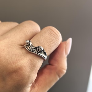 Op de afbeelding: Een zilveren kikkerring. De ring heeft een gedetailleerd kikkerontwerp met prominente ogen en een getextureerde huid. Het lichaam van de kikker wikkelt zich om de vinger en creëert een uniek accessoire. De ring wordt op een vinger getoond.
