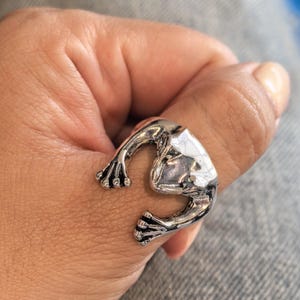 Könnte beinhalten: Ein silberfarbener Metallring in Form eines Frosches, der sich um den Finger legt. Das Design zeigt einen detaillierten Froschkopf und Vorderbeine mit sichtbaren Zehen, abgerundet durch eine polierte, reflektierende Oberfläche und abgedunkelte vertiefte Bereiche für Kontraste.