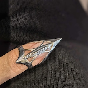 Puede incluir: Un anillo de dedo plateado con un diseño afilado y puntiagudo. El anillo presenta un elemento alargado en forma de punta de flecha que se extiende sobre la yema del dedo, con detalles intrincados. El anillo se lleva en un dedo.