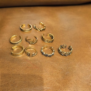 Puede incluir: Una colección de ocho anillos de metal en tono dorado, cada uno con un diseño abierto. El conjunto incluye varias texturas y patrones, como bandas lisas, detalles de cuerda trenzada, formas onduladas y diseños intrincados con recortes.