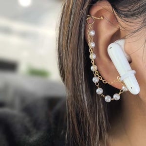 May include: Gold-coloured ear cuff featuring a chain of faux pearls and a white earbud holder. The ear cuff wraps around the ear and connects to the earbud holder with a delicate chain. The pearls are small and round.