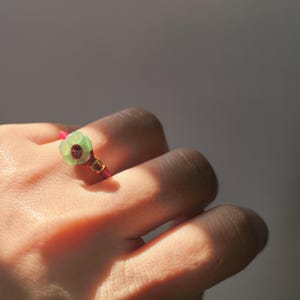 May include: A close-up of a ring featuring a light green flower-shaped gemstone with a red center. The ring's band is a deep red cord, accented with gold-colored details. The ring is on a person's finger.