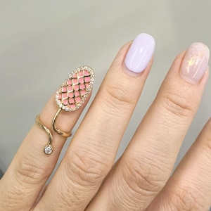 Può includere: Un anello dorato con un design a forma di serpente, con una parte superiore rosa a motivo a griglia incastonata con piccole pietre trasparenti. Una piccola pietra trasparente accentua la coda del serpente. L'anello è indossato su un dito con smalto lilla chiaro.