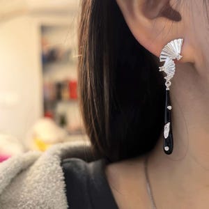 May include: Silver-toned fan-shaped earrings with a black teardrop-shaped dangle. The earrings feature small, clear gemstones and are worn on the ear. The fan design has a textured, layered appearance.