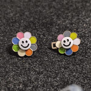 May include: Two flower-shaped enamel pins with a smiling face in the center. The petals are a mix of colors including pink, blue, green, orange, and gray. One pin has a clasp on the back.