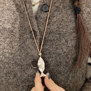 May include: A silver and gold fish pendant necklace with a beige cord. The fish pendant has intricate detailing and is held by a person. The background is a brown wool sweater with dark brown buttons.