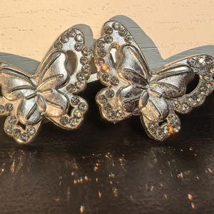 May include: Two silver-toned butterfly-shaped decorative objects. Each butterfly has intricate wing details and is embellished with small, clear rhinestones. The butterflies are displayed on a dark, reflective surface.