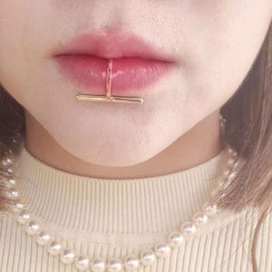 May include: A gold-toned metal lip cuff featuring a horizontal bar design that rests against the lower lip. The jewellery piece is worn vertically, with a thin band extending from the bar to hook over the lip, paired with a white pearl necklace.