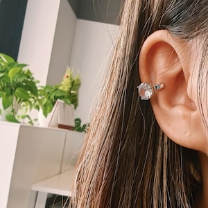 May include: A silver ear cuff with a large, clear, round gemstone. The cuff wraps around the ear, giving the impression of a ring. The jewellery is set against a backdrop of brown hair and a blurred interior.