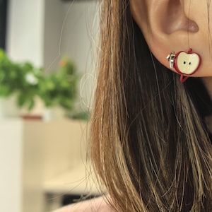 May include: Red and white apple-shaped earrings. The earrings feature a red outline with a white center and two black dots. The earrings are attached to a silver-colored clasp. The earrings are worn on a person's ear.