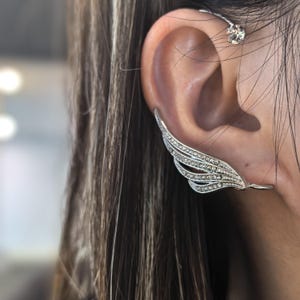 May include: A silver ear cuff with a wing design, adorned with small, sparkling crystals. The cuff curves along the ear, with a separate stud earring visible at the top. The jewelry is set against a blurred background.