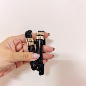 May include: Black hair ties with gold-colored metal accents and clear crystal embellishments. The hair ties are made of a woven material and are held in a hand against a white background.