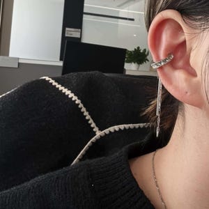 May include: Silver ear cuff with a row of clear crystals and dangling chain detail. The ear cuff wraps around the upper ear and features a delicate, dangling chain that adds movement and sparkle. A stylish accessory for any occasion.